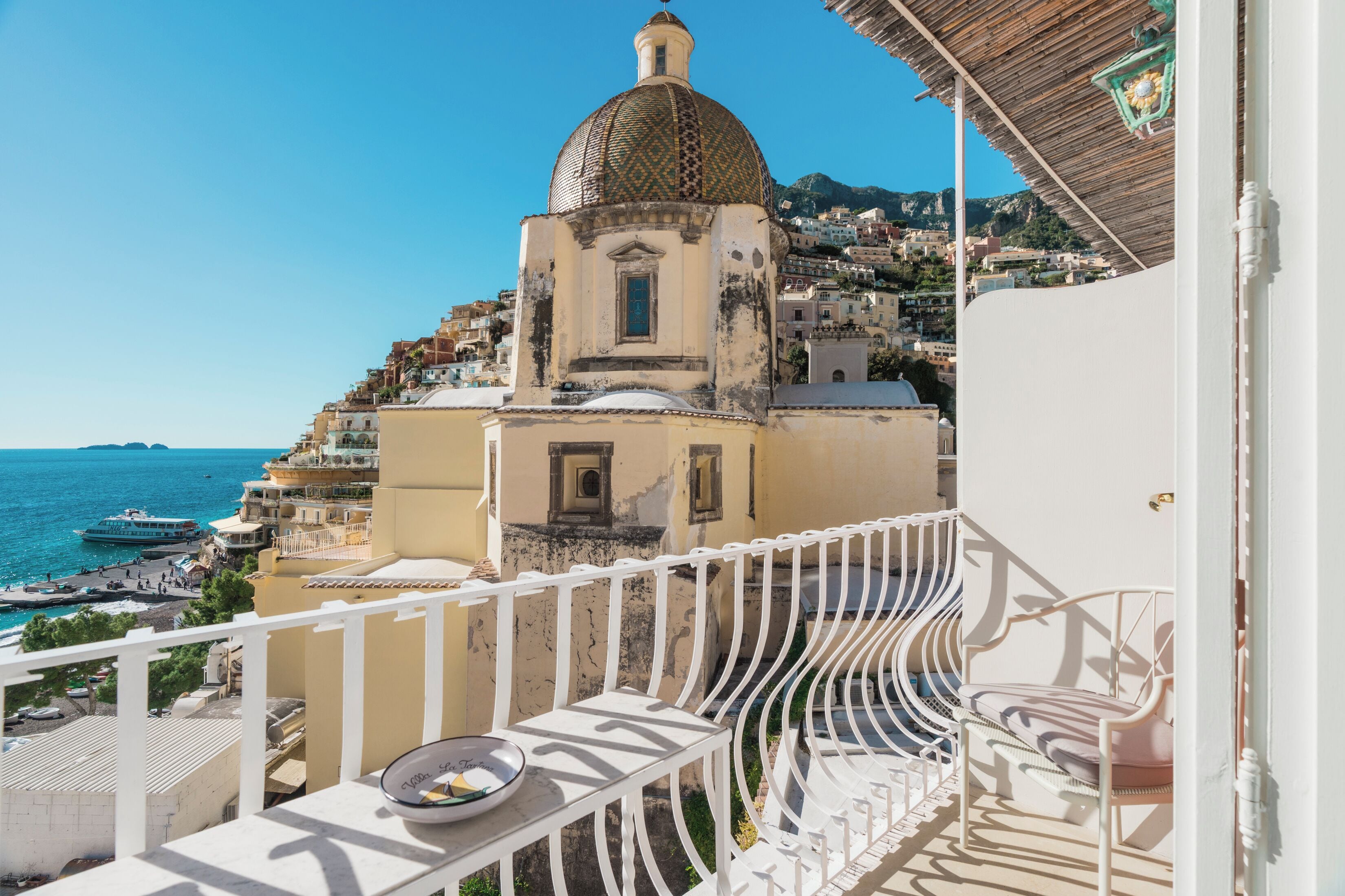 Un balcon avec une balustrade blanche donnant sur une ville côtière, un bâtiment surmonté d’un dôme et un ciel bleu dégagé.