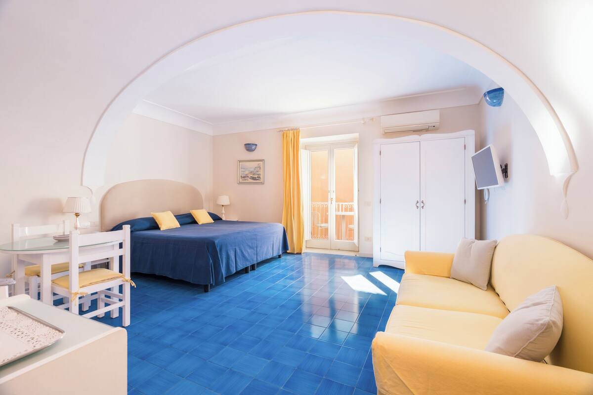 A hotel room with a bed, a sofa, a desk with a lamp, and a TV.