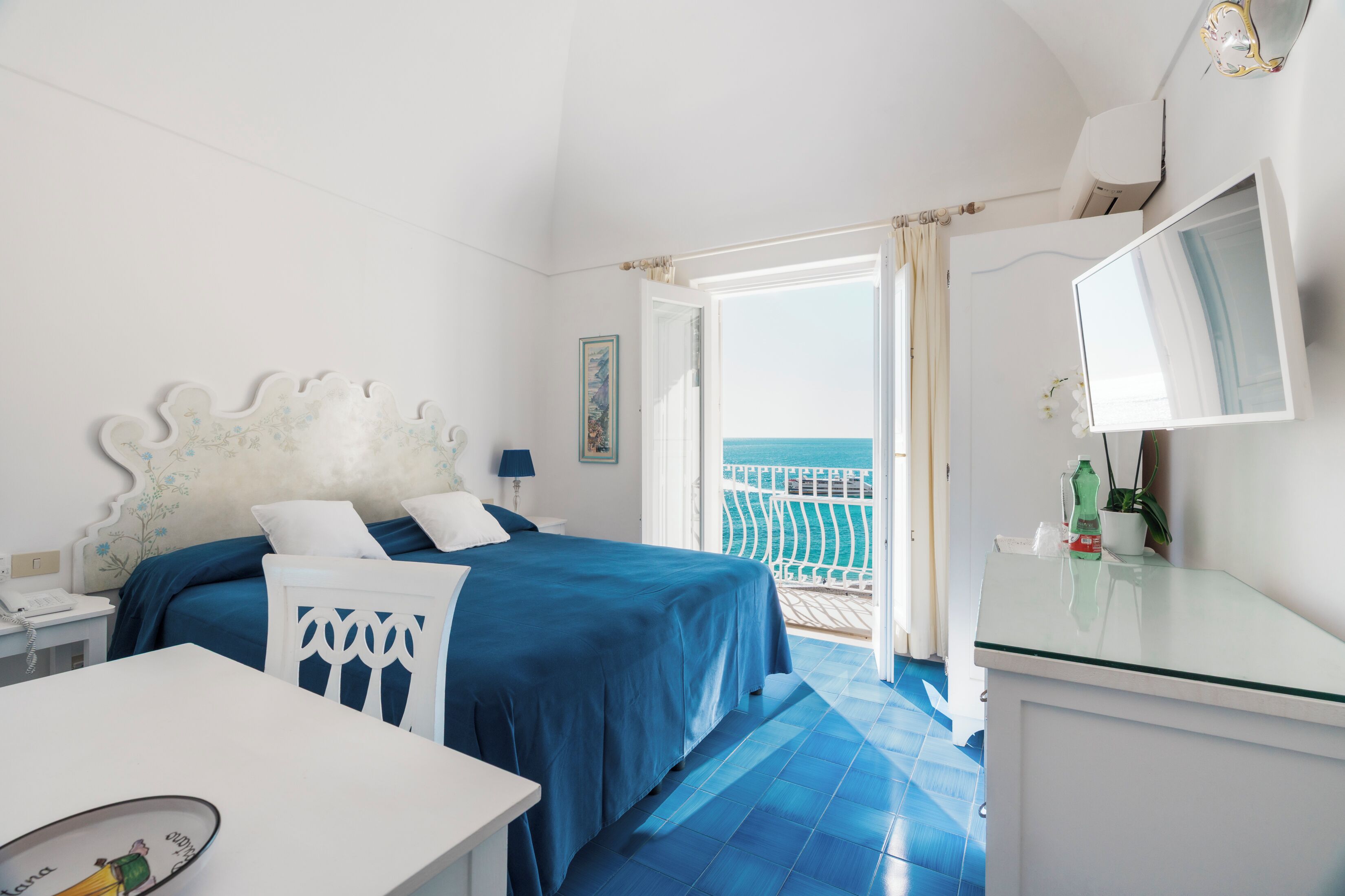A bedroom with a bed, a desk, and a balcony with a view of the sea.