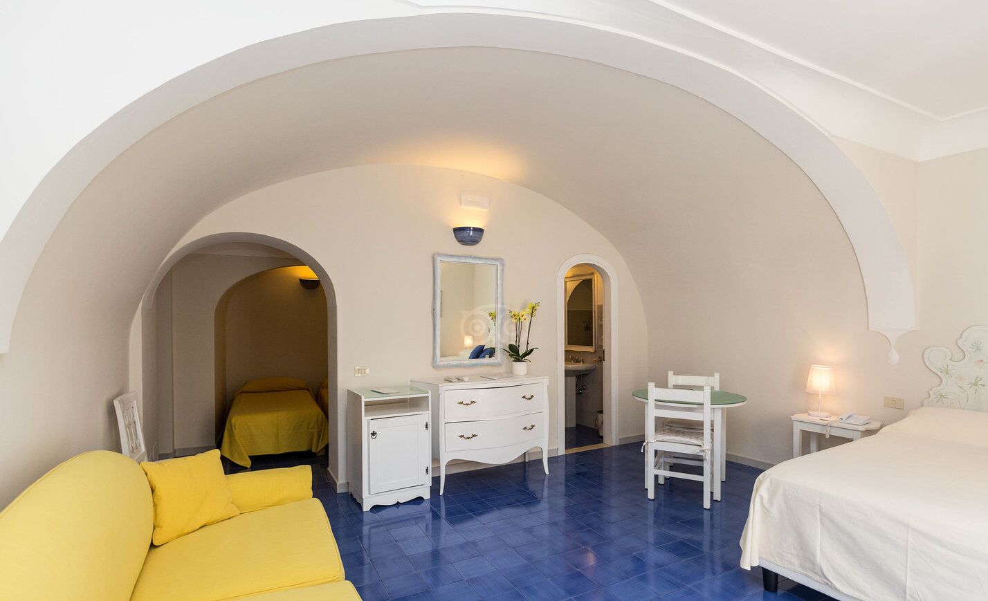A hotel room with a bed, a desk, a chair, and a small table. There is a mirror above the desk and a window with a view of the outside.