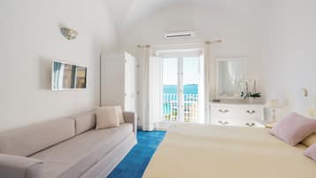 A bedroom with a bed, a sofa, a dresser, and a large window with a view of the sea.