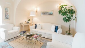 Un salón con muebles blancos, una mesa de centro de cristal con libros, una lámpara y una planta.