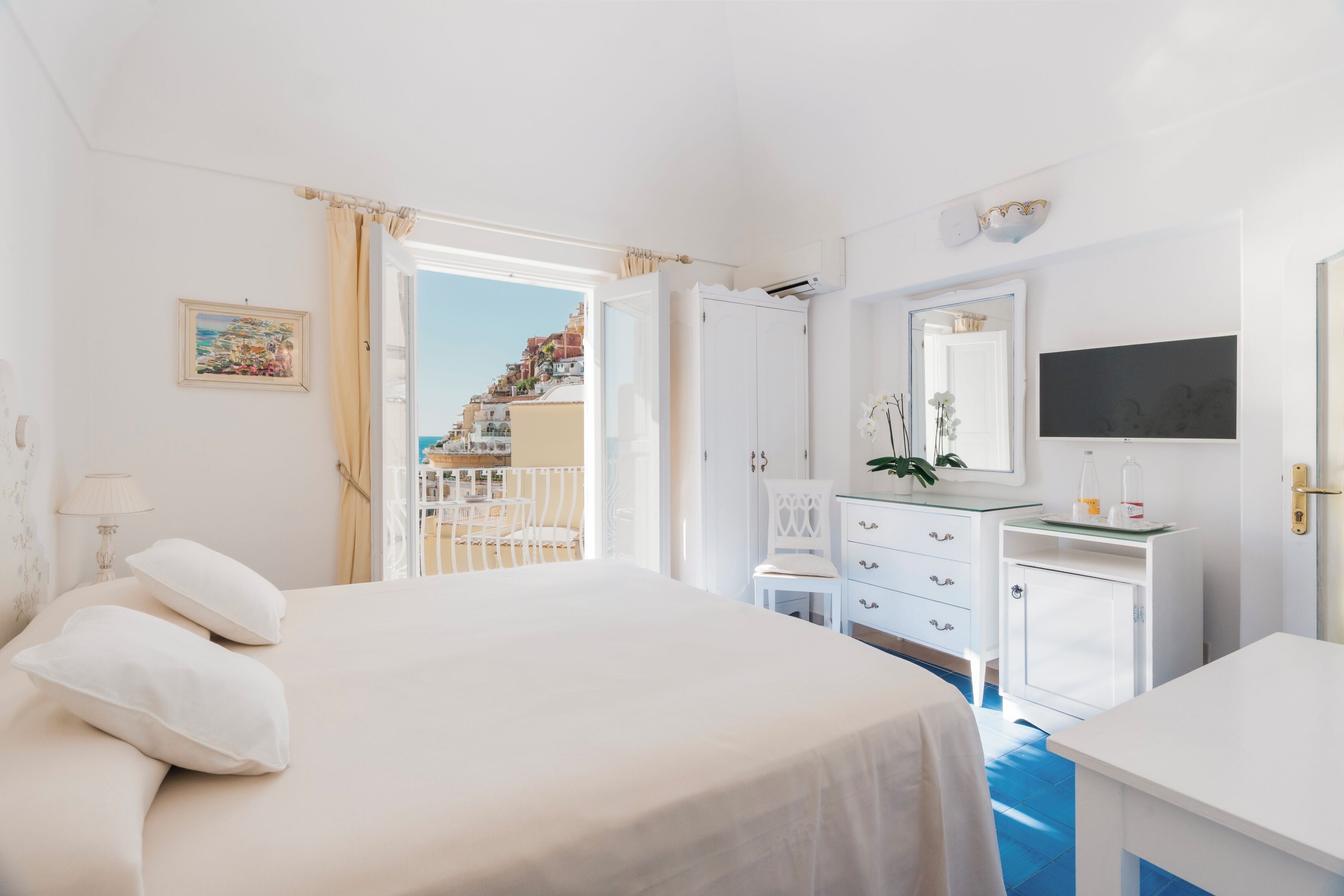 A hotel room with a bed, a desk, a dresser, and a view of a coastal town.