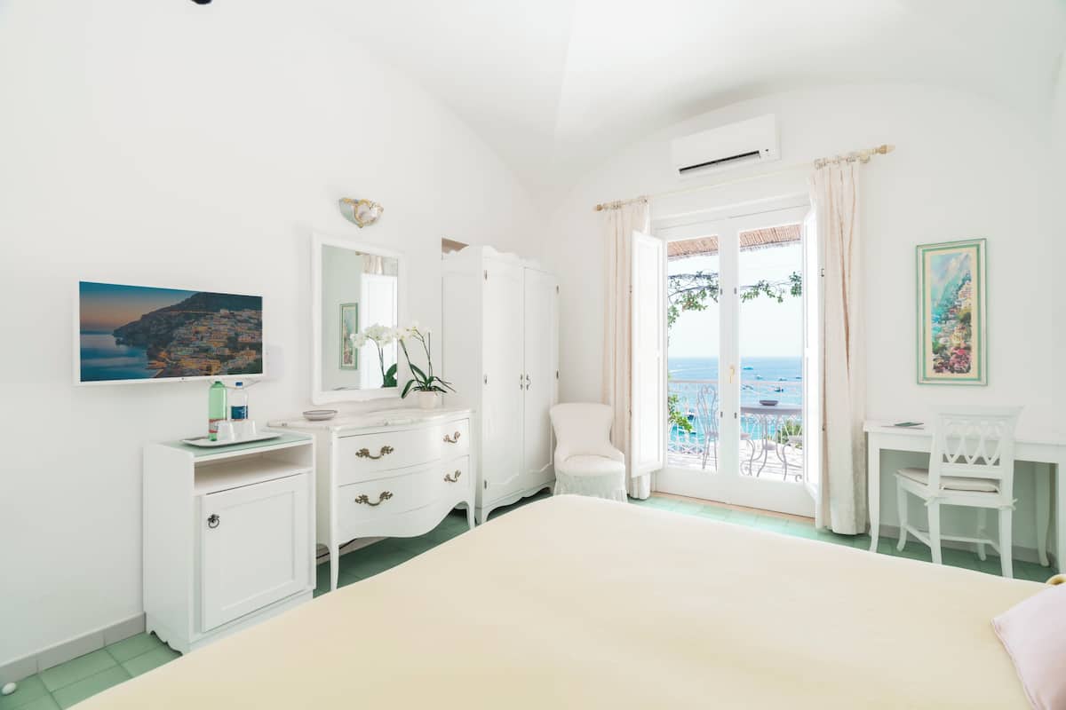 A bedroom with a bed, a dresser, a chair, a desk, and a balcony with a view of the sea.