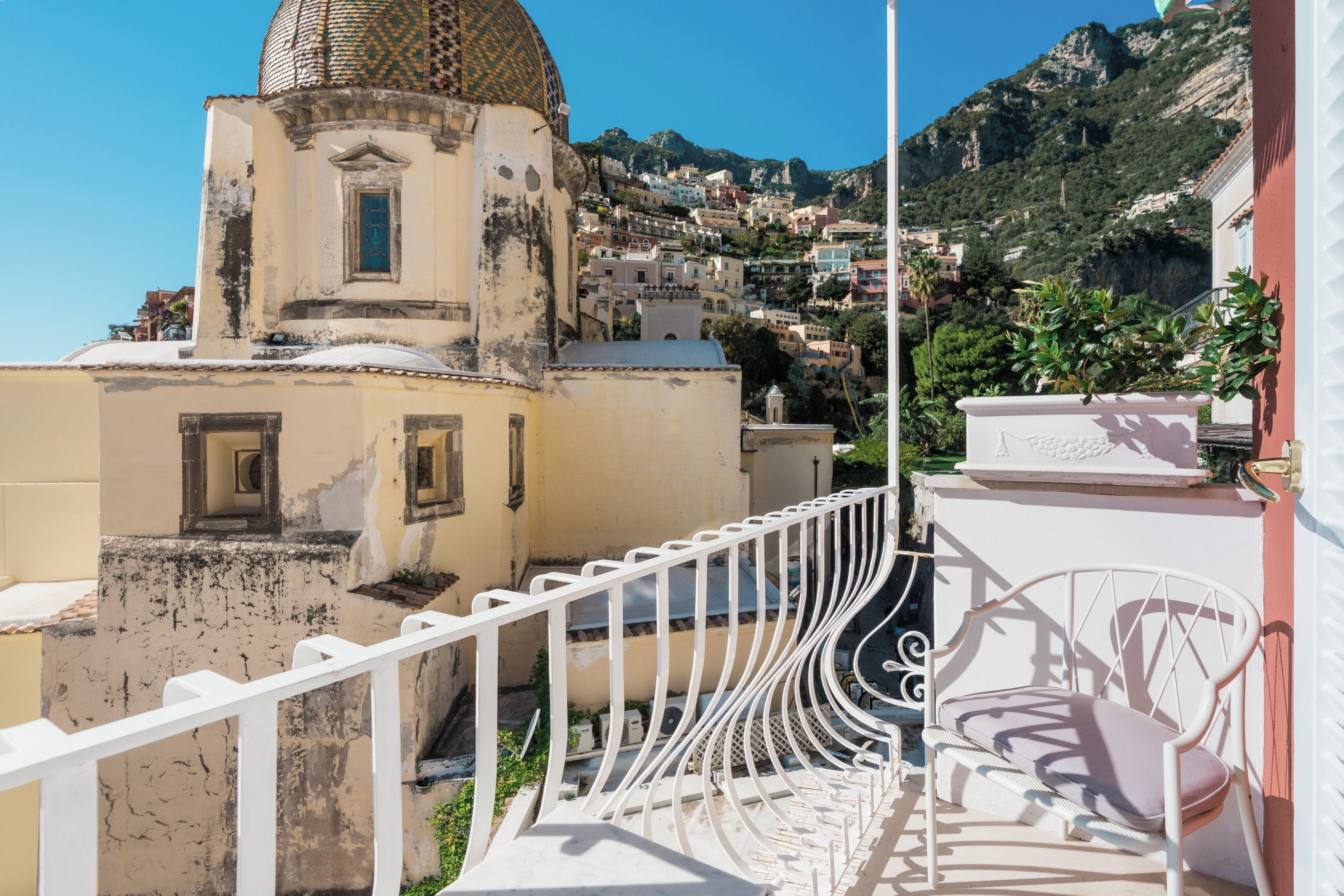 A balcony with a white railing, a white chair, and a view of a hillside with buildings.