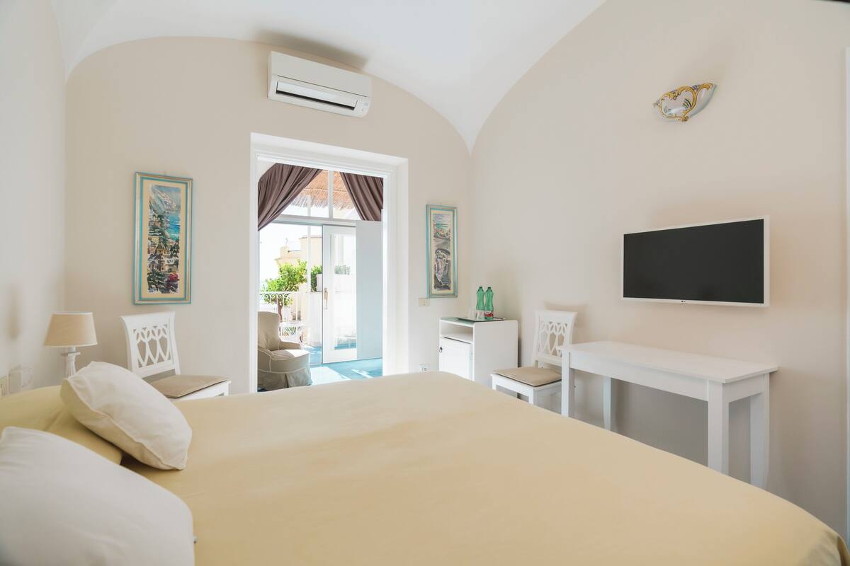 A hotel room with a large bed, a desk, a chair, a television, and a door leading to a balcony.
