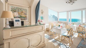 Un vestíbulo de hotel con recepción, comedor con mesas y sillas, amplias ventanas con vistas al mar y obras de arte en las paredes.