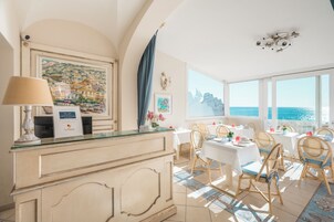 Un vestíbulo de hotel con recepción, comedor con mesas y sillas, amplias ventanas con vistas al mar y obras de arte en las paredes.