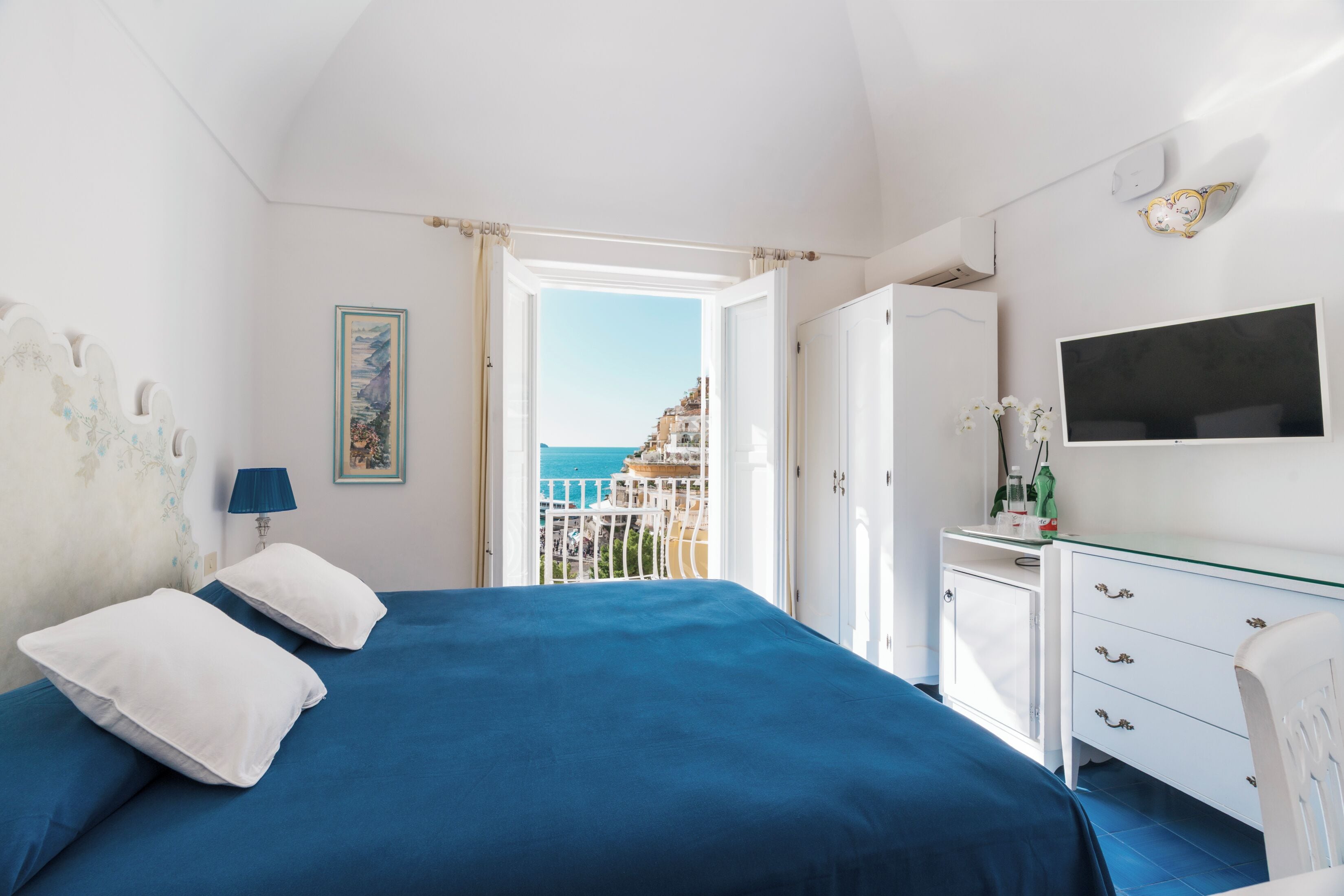 Une chambre à coucher comprenant un lit, une commode, un téléviseur et donnant sur un balcon avec vue sur la ville.