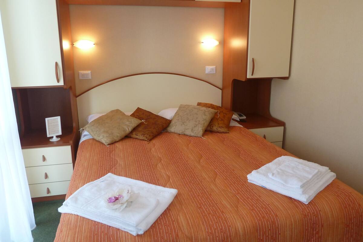 A neatly made bed with a striped bedspread, two pillows, and a folded towel with a flower. There are bedside tables with lamps and a small screen.