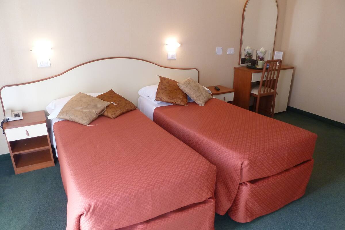 A double room with two beds, each with a red bedspread and matching pillows. There is a wooden nightstand with a phone and a mirror on the wall.