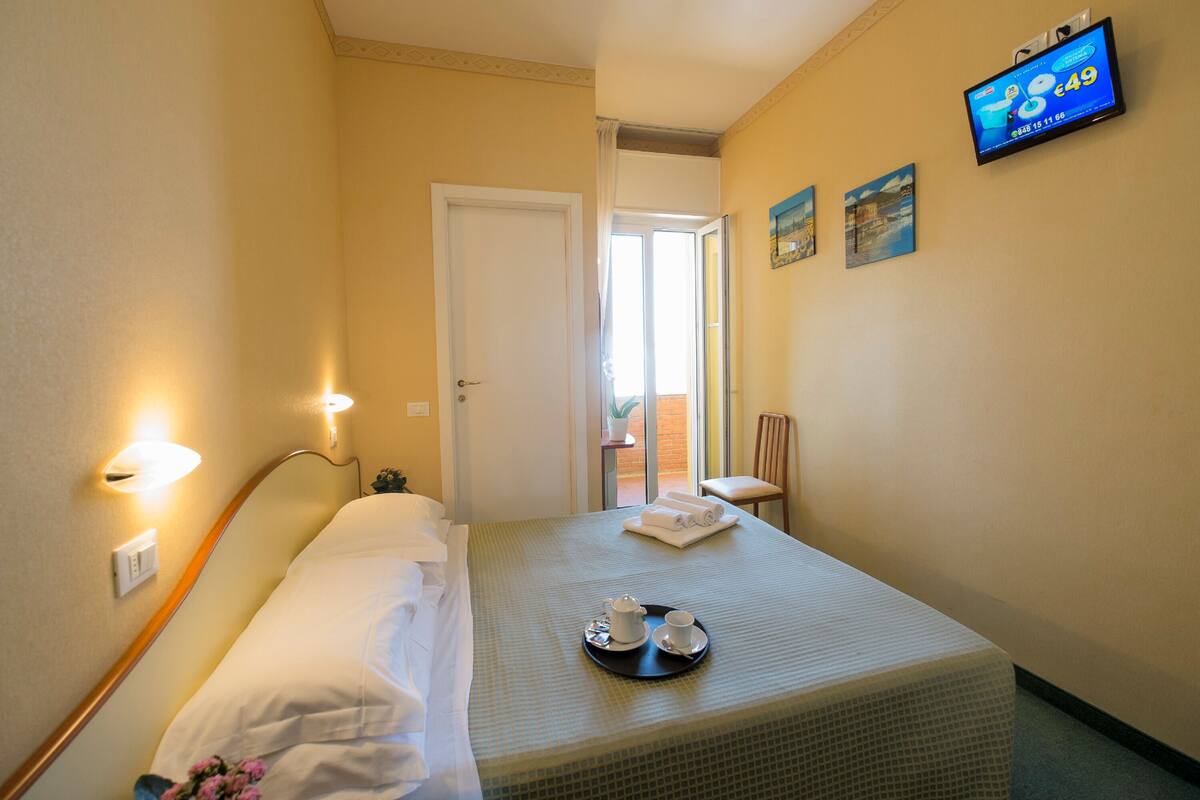 A hotel room with a bed, a tray with a teapot and cups, a television on the wall, and a door leading to a balcony.