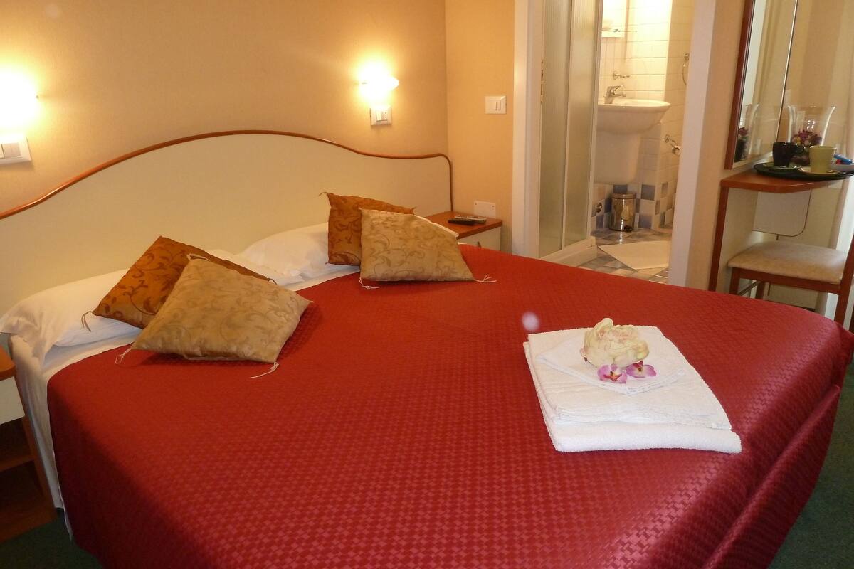 A hotel room with a large bed, two pillows, a nightstand, a mirror, and a bathroom visible through a glass door.