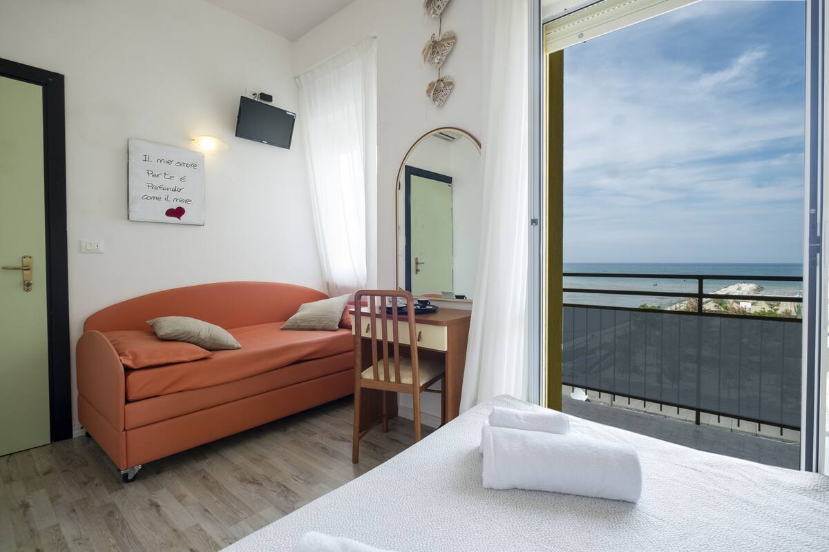 A bedroom with a bed, a desk, a chair, a sofa, a window with a view of the sea, and a wall with a framed quote.