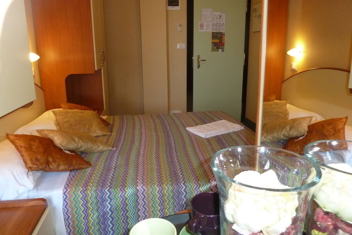 A small, cozy room with a bed, pillows, a glass vase with flowers, and a door with a notice.