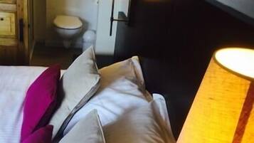 A hotel room with a bed, pillows, a nightstand, a lamp, a wooden cabinet, and a toilet.