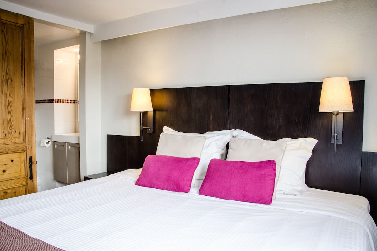 Une chambre d’hôtel avec un grand lit, deux lampes de chevet, une armoire en bois et une petite salle de bain équipée de toilettes et d’un lavabo.