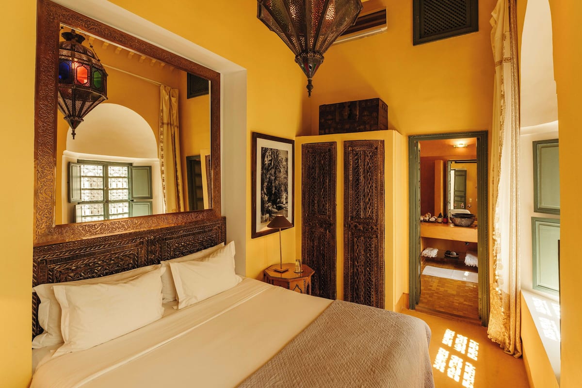 A hotel room with a large bed, a mirror, a wooden headboard, a small table with a lamp, and a wardrobe with a mirror.