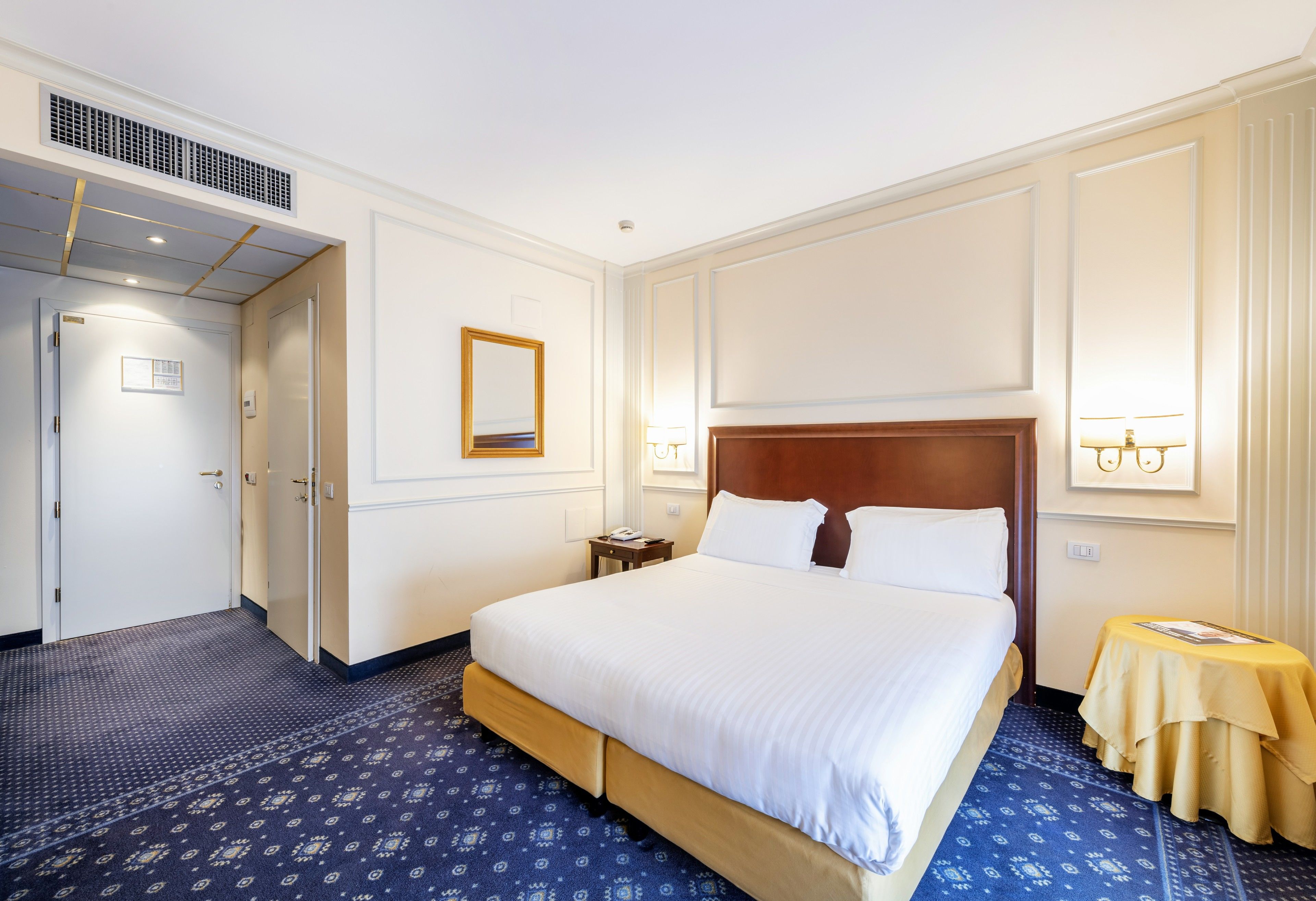 Hotelkamer met een groot bed, nachtkastje, spiegel en een wandlamp.