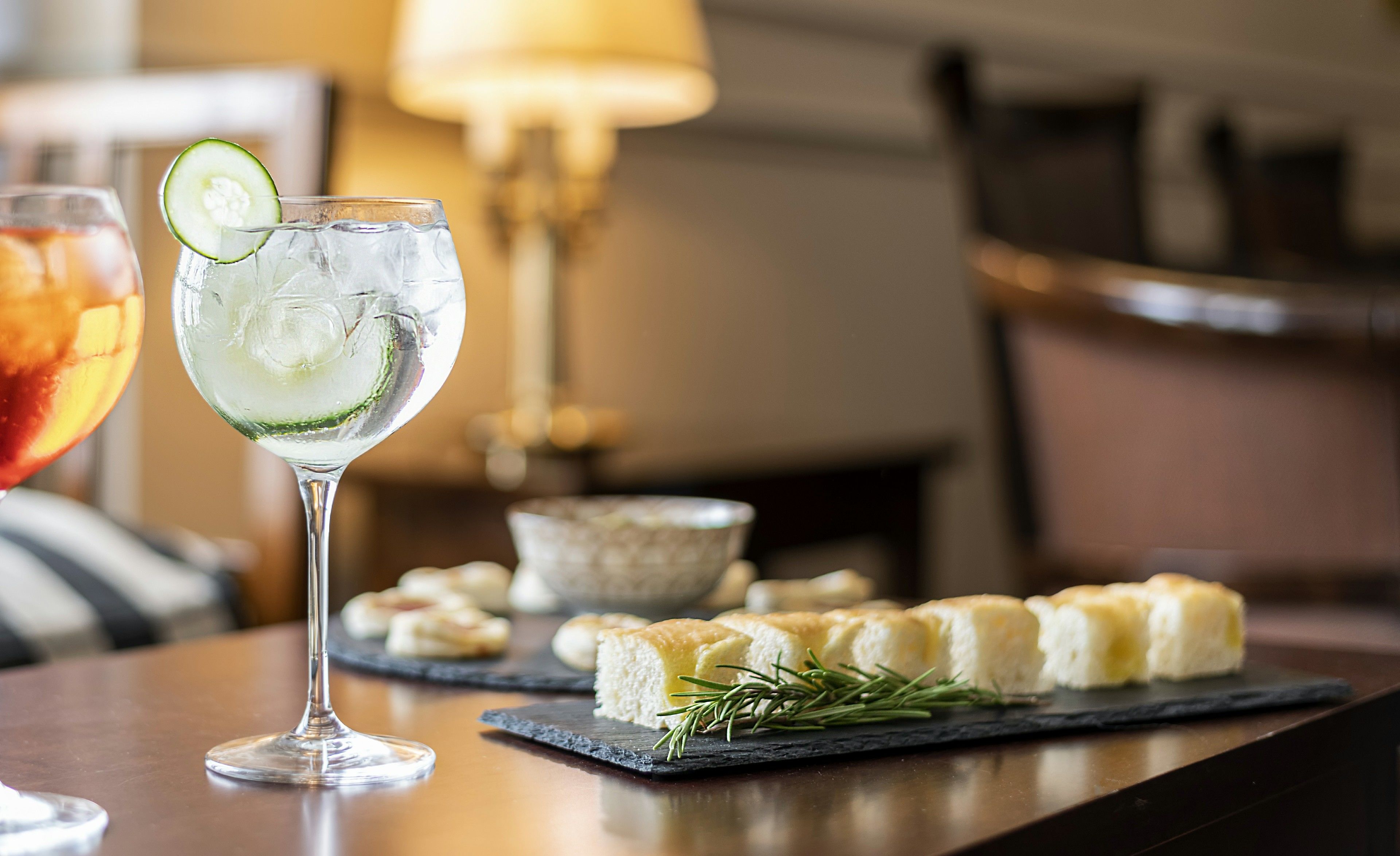 A bar setting with a glass of a clear beverage garnished with a slice of cucumber, a glass of red drink, and a slate board with pastries and rosemary.