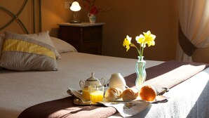 A neatly made bed with pillows, a bedside table with a lamp and vase, and a breakfast tray with croissants, orange, and a vase with flowers. - B&B Camere Andrei (Pienza)