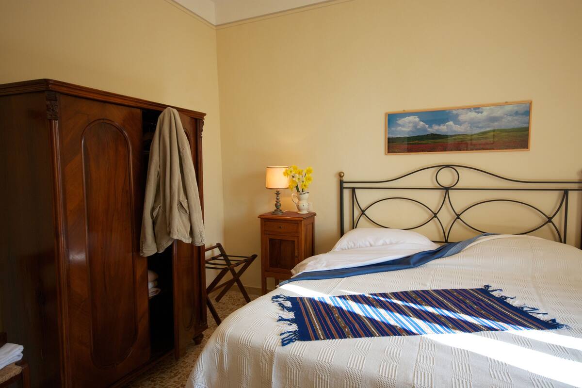 A bedroom with a bed, a wooden wardrobe, a nightstand with a lamp, and a painting on the wall.