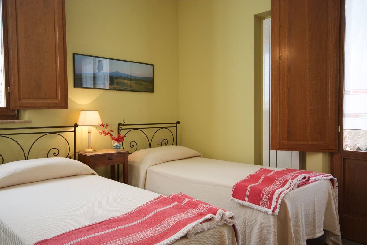 A room with two beds, a nightstand with a lamp, a vase with flowers, and a painting on the wall.