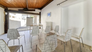Una habitación con techo de madera, paredes blancas y vistas a edificios. Seis sillas con diseños geométricos y una mesa con superficie blanca.
