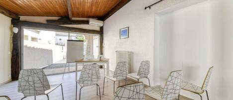 A room with a wooden ceiling, white walls, and a view of buildings outside. There are six chairs with geometric patterns and a table with a white surface.
