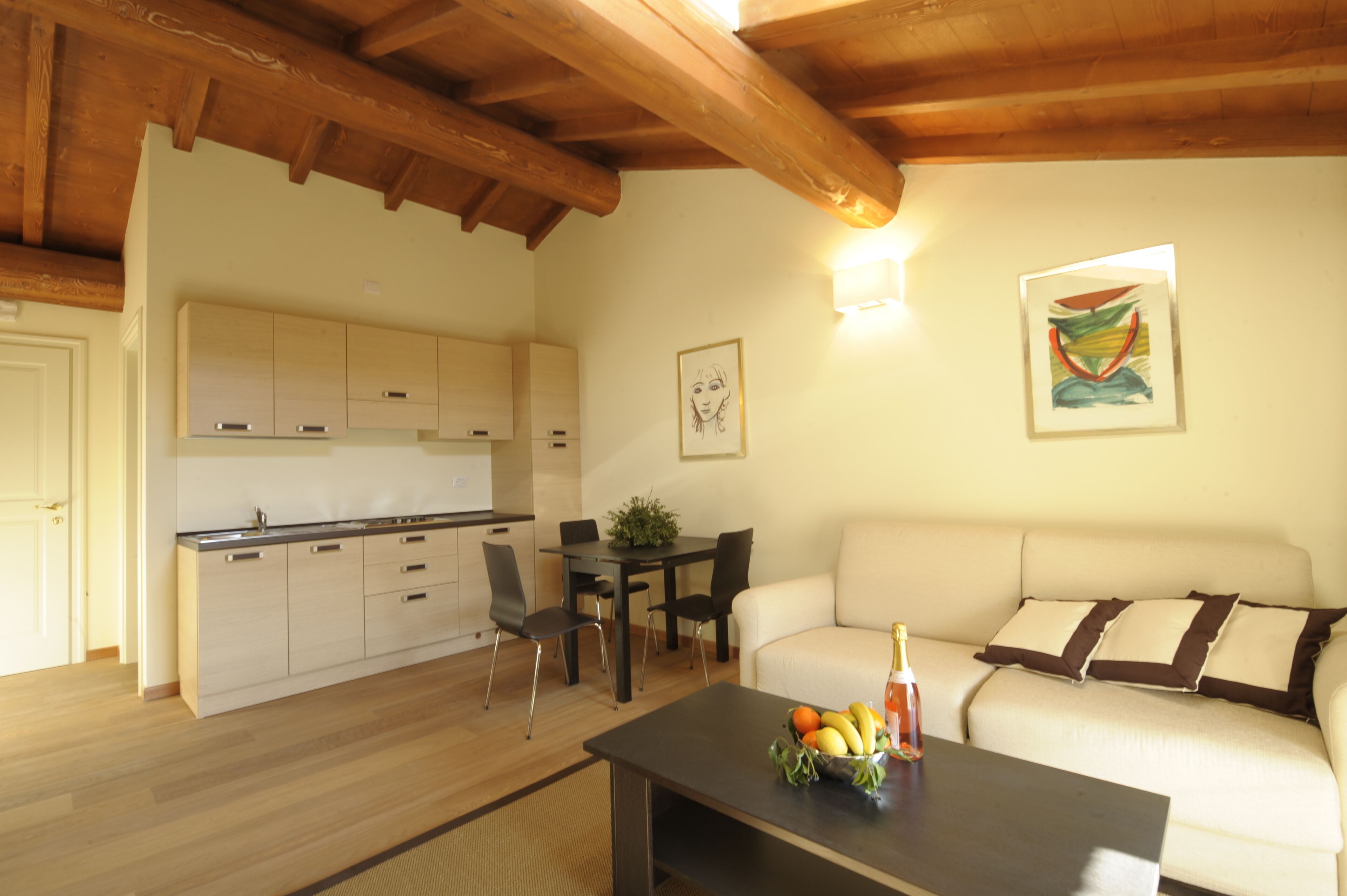 Une cuisine moderne avec des poutres en bois, un coin repas avec une table et des chaises, un salon avec un canapé et une petite table sur laquelle sont disposés des fruits et une bouteille.