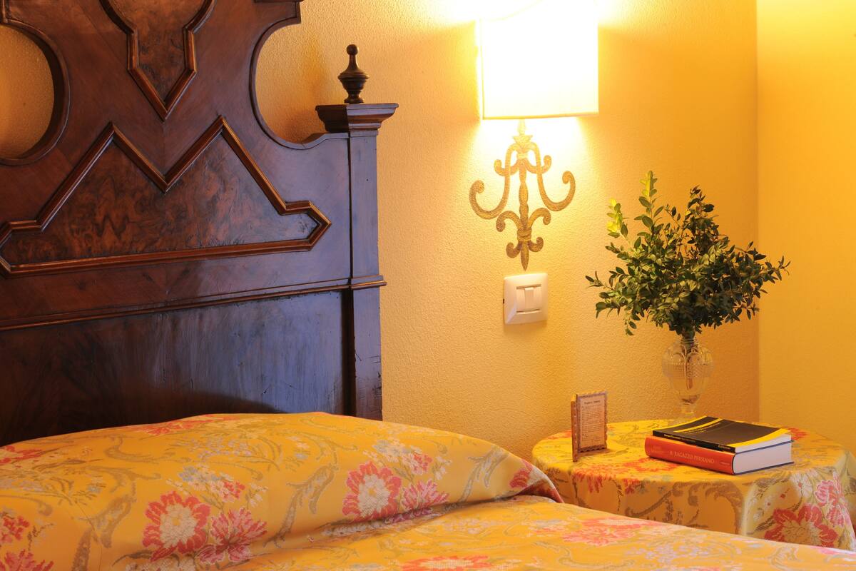 A hotel room with a wooden headboard, a bedside table with a lamp, a potted plant, and a book on the table.
