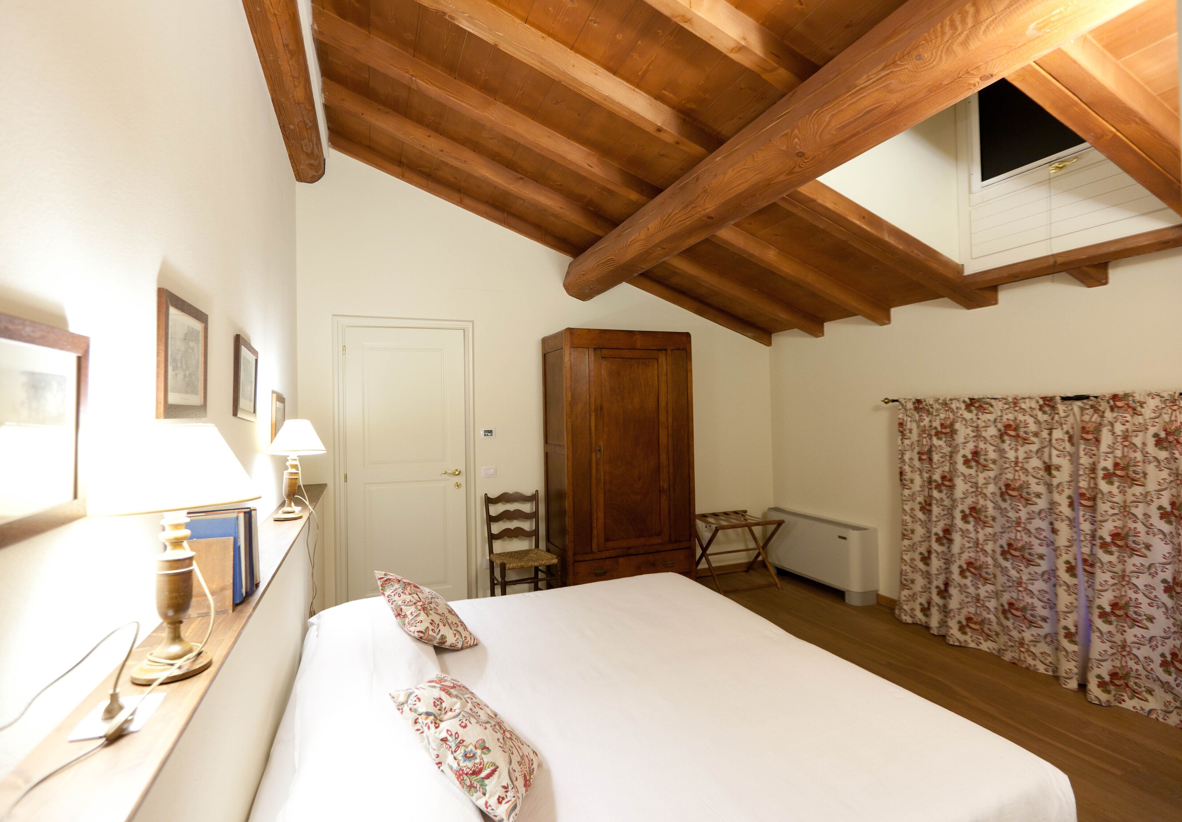 Un dormitorio con techo de madera, una cama con ropa de cama floral, un armario de madera, una silla, un escritorio con una lámpara y una ventana con cortinas.