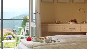Un dormitorio con cama, una mesita de noche con un frutero, una taza y una tetera. Hay tres cuadros en la pared y una ventana con vistas a la vegetación y montañas.