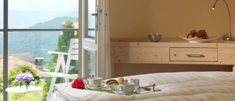 Ein Schlafzimmer mit einem Bett, einem Nachttisch mit einer Obstschale, einer Tasse und einem Teekessel. An der Wand hängen drei gerahmte Bilder und ein Fenster mit Blick auf Grünflächen und Berge.