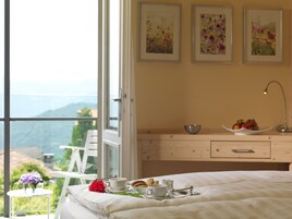 Un dormitorio con cama, una mesita de noche con un frutero, una taza y una tetera. Hay tres cuadros en la pared y una ventana con vistas a la vegetación y montañas.