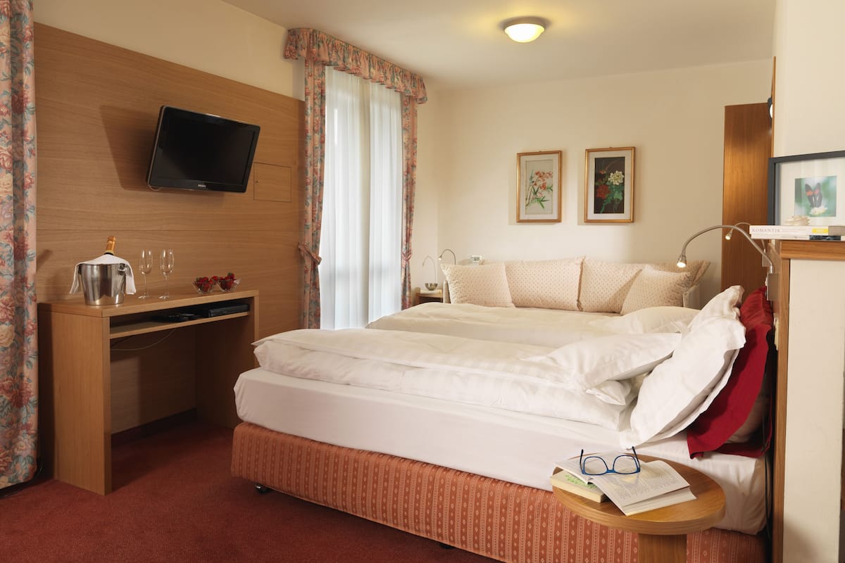 Habitación de hotel con cama, escritorio con televisor, mesita de noche con vasos y un libro, y dos cuadros en la pared.