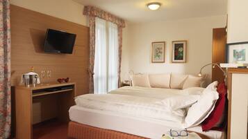 A hotel room with a bed, a desk with a TV, a bedside table with glasses and a book, and two framed pictures on the wall.