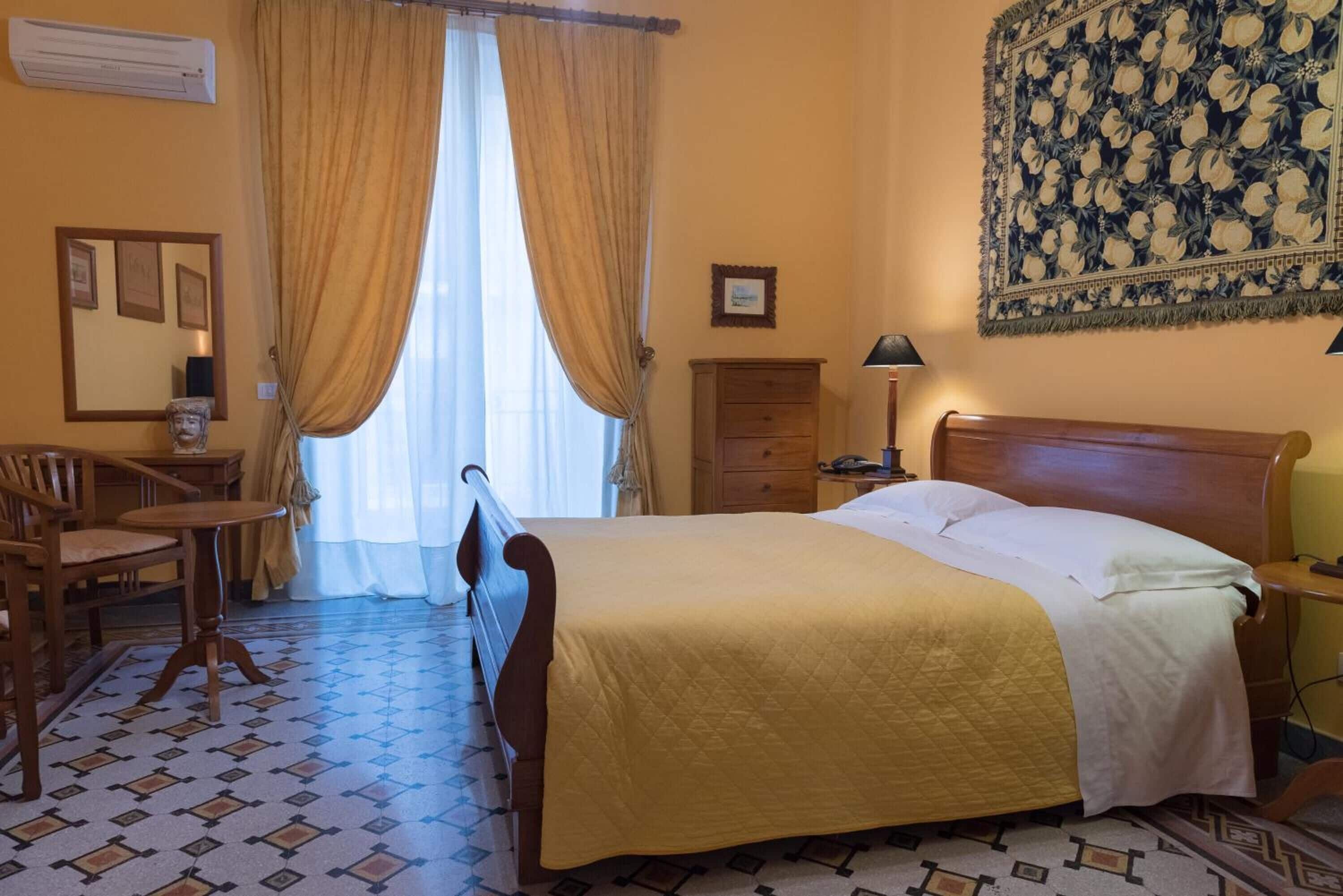 A hotel room with a bed, a desk, a chair, a nightstand, a lamp, and a tapestry on the wall.