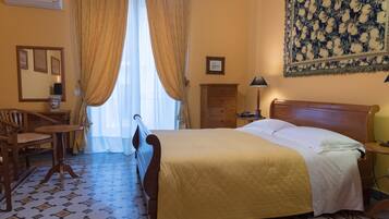 A hotel room with a bed, a desk, a chair, a nightstand, a lamp, and a tapestry on the wall.
