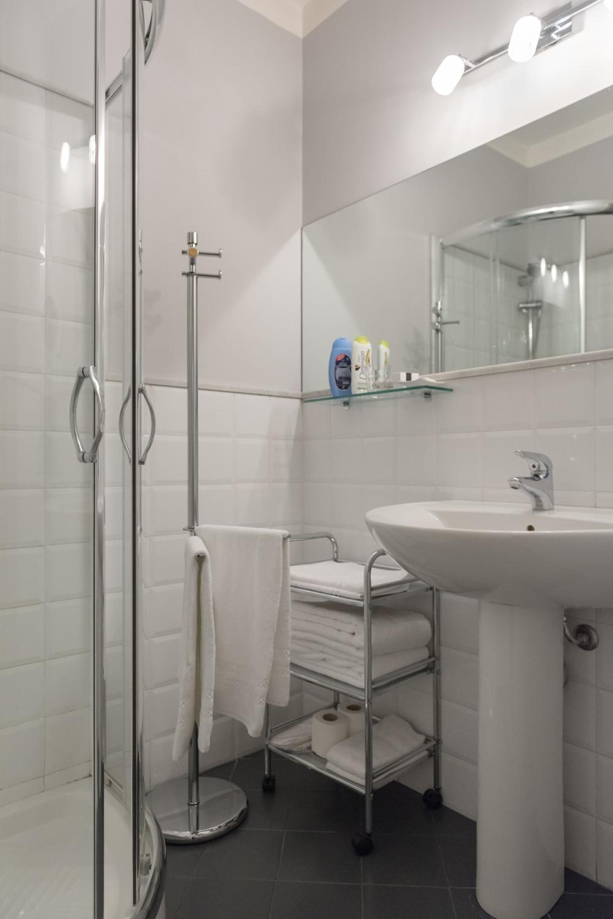 Un baño moderno con cabina de ducha de vidrio, un lavabo blanco y un espejo encima. Hay toallas y artículos de aseo en un estante.