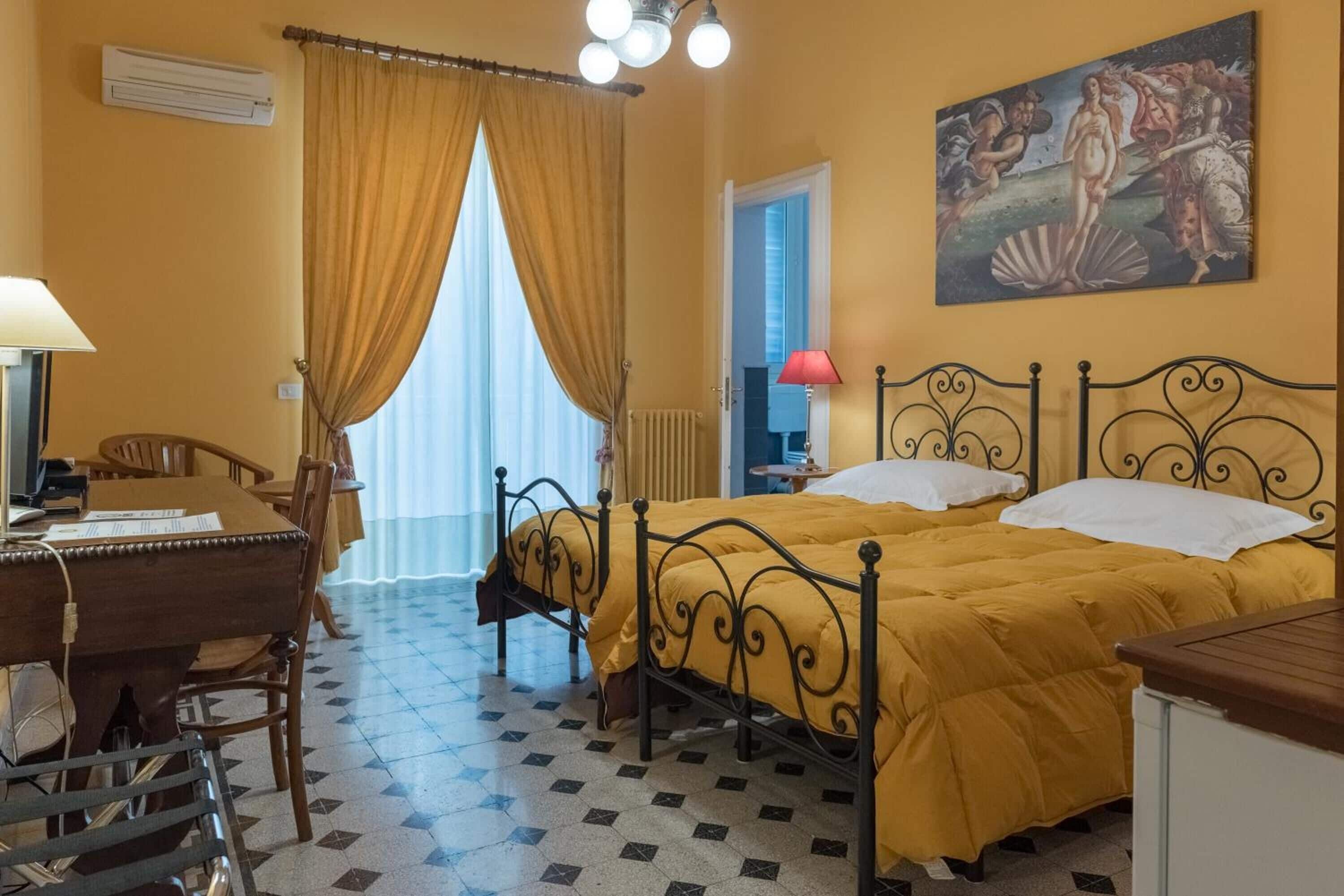 Una stanza con un letto, una scrivania e una sedia. Si vede una finestra con tende, un termosifone e un quadro appeso al muro.