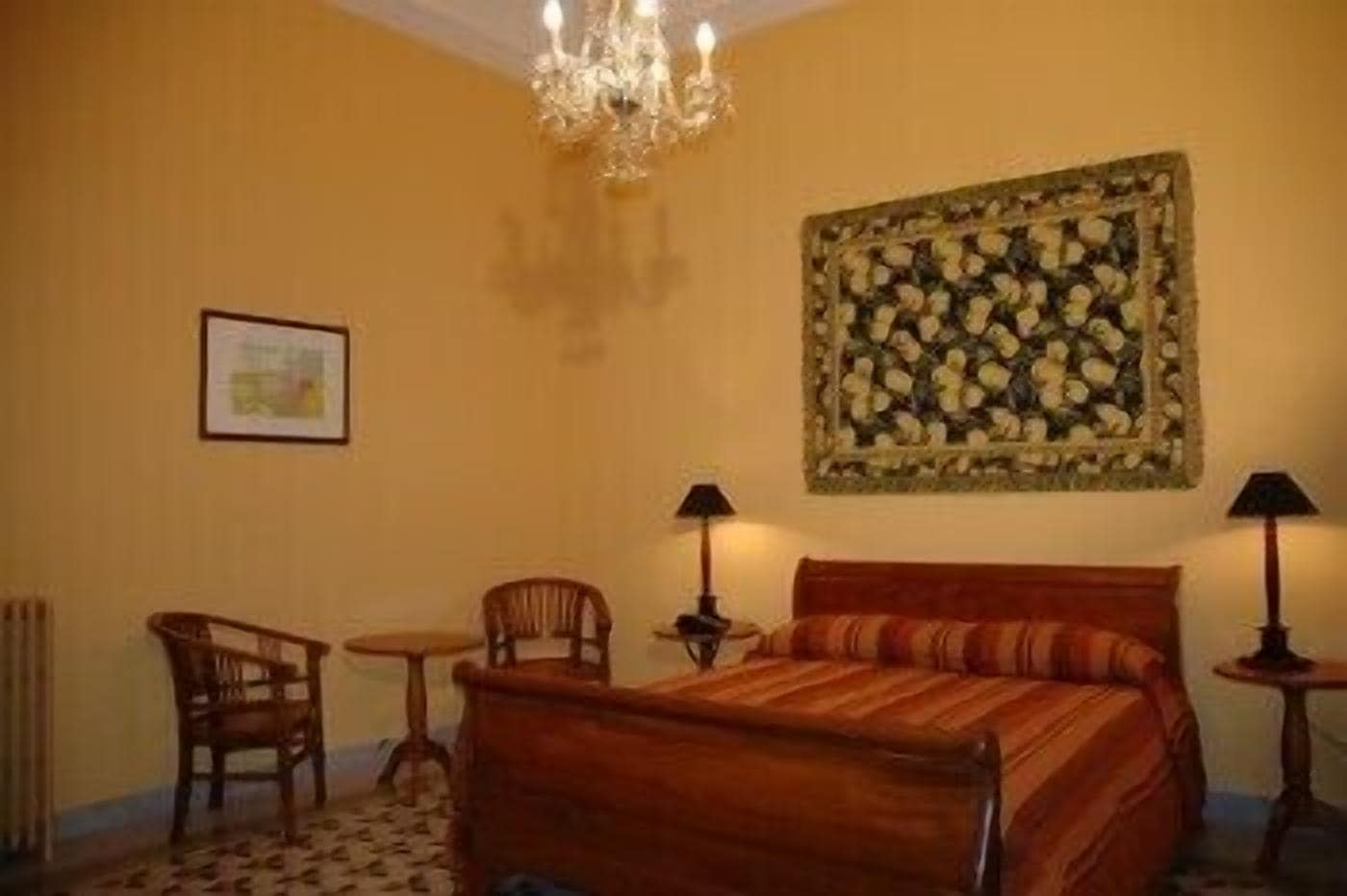 Una camera da letto con un letto di legno, due sedie, un tavolino, un lampadario e un arazzo floreale sulla parete.