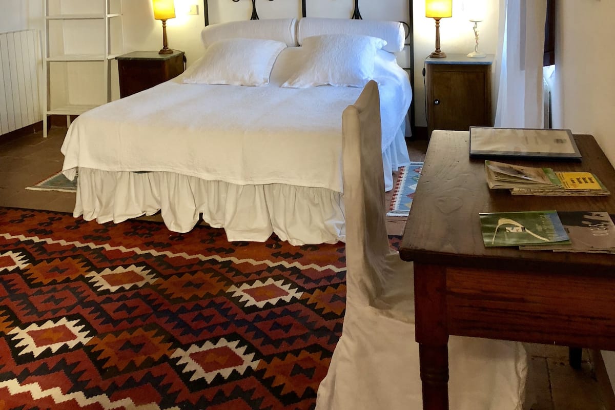 Un dormitorio con una cama, dos mesitas de noche con lámparas, un escritorio de madera, una alfombra estampada y una escalera apoyada en la pared.