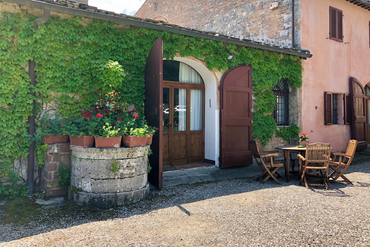 Un edificio rústico con fachada de piedra parcialmente cubierta de hiedra, una puerta de madera y un patio de grava con muebles de madera.