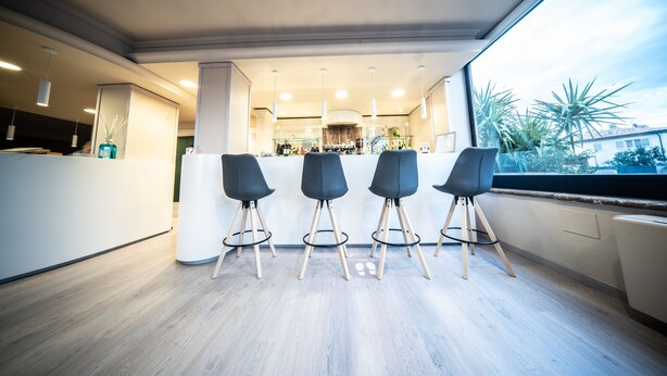 A modern kitchen with a bar area, featuring three bar stools, a large window with a view of palm trees, and a well-lit interior. - Hotel Le Grazie (Assisi)