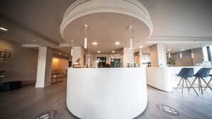 A modern reception area with a central white counter, wooden flooring, and a staircase in the background. - Hotel Le Grazie (Assisi)