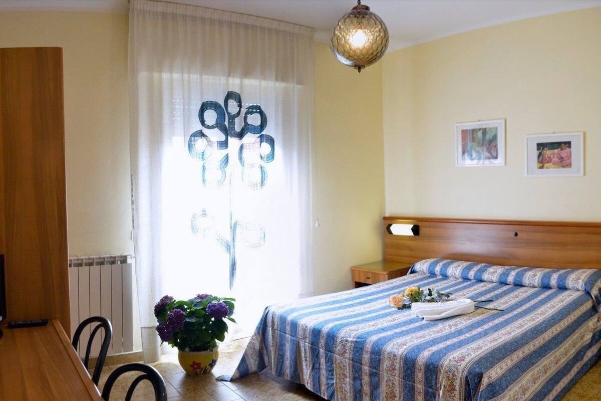 A bedroom with a bed, a wooden desk, a potted plant, and a window with curtains.