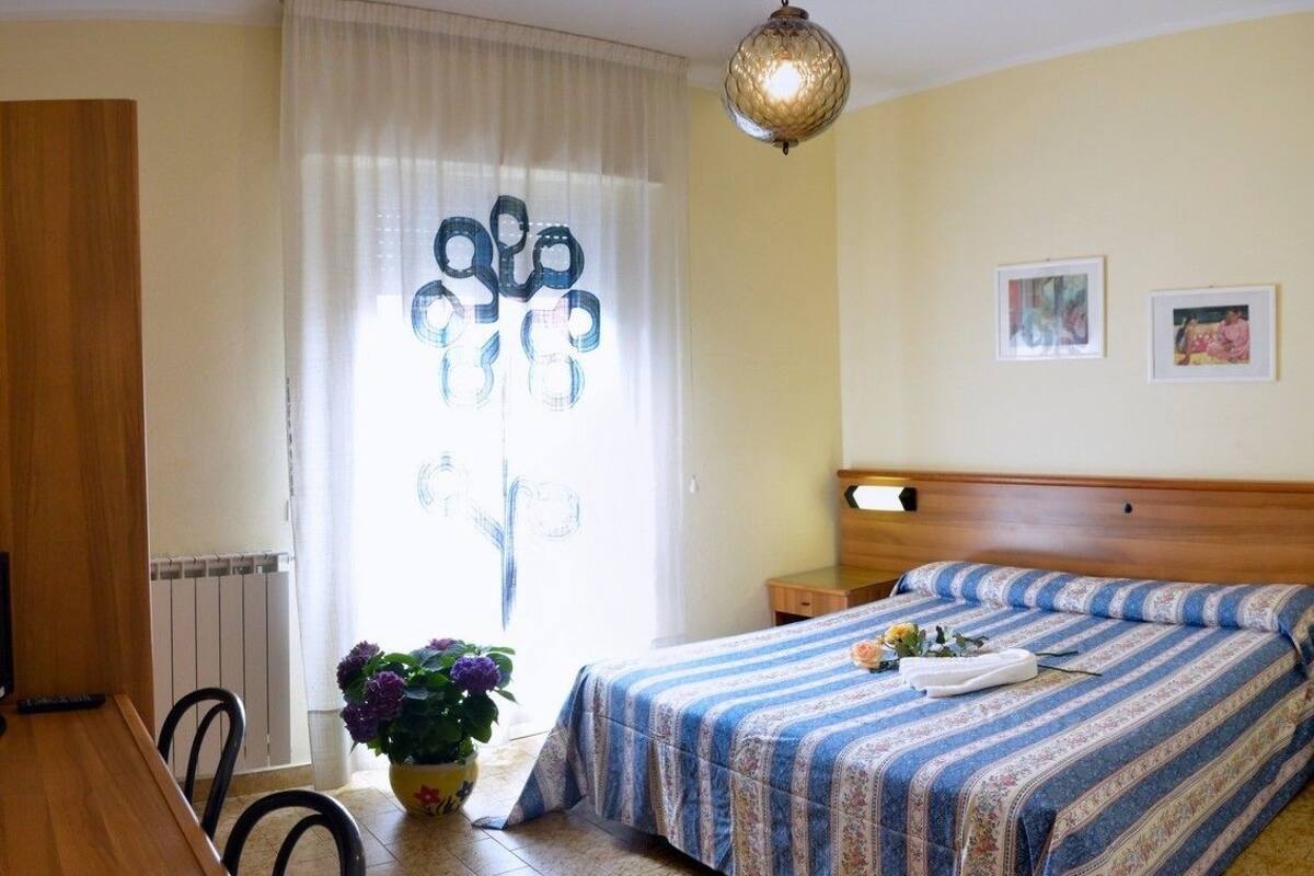 A hotel room with a bed, a wooden desk, a chair, a potted plant, and a window with curtains.