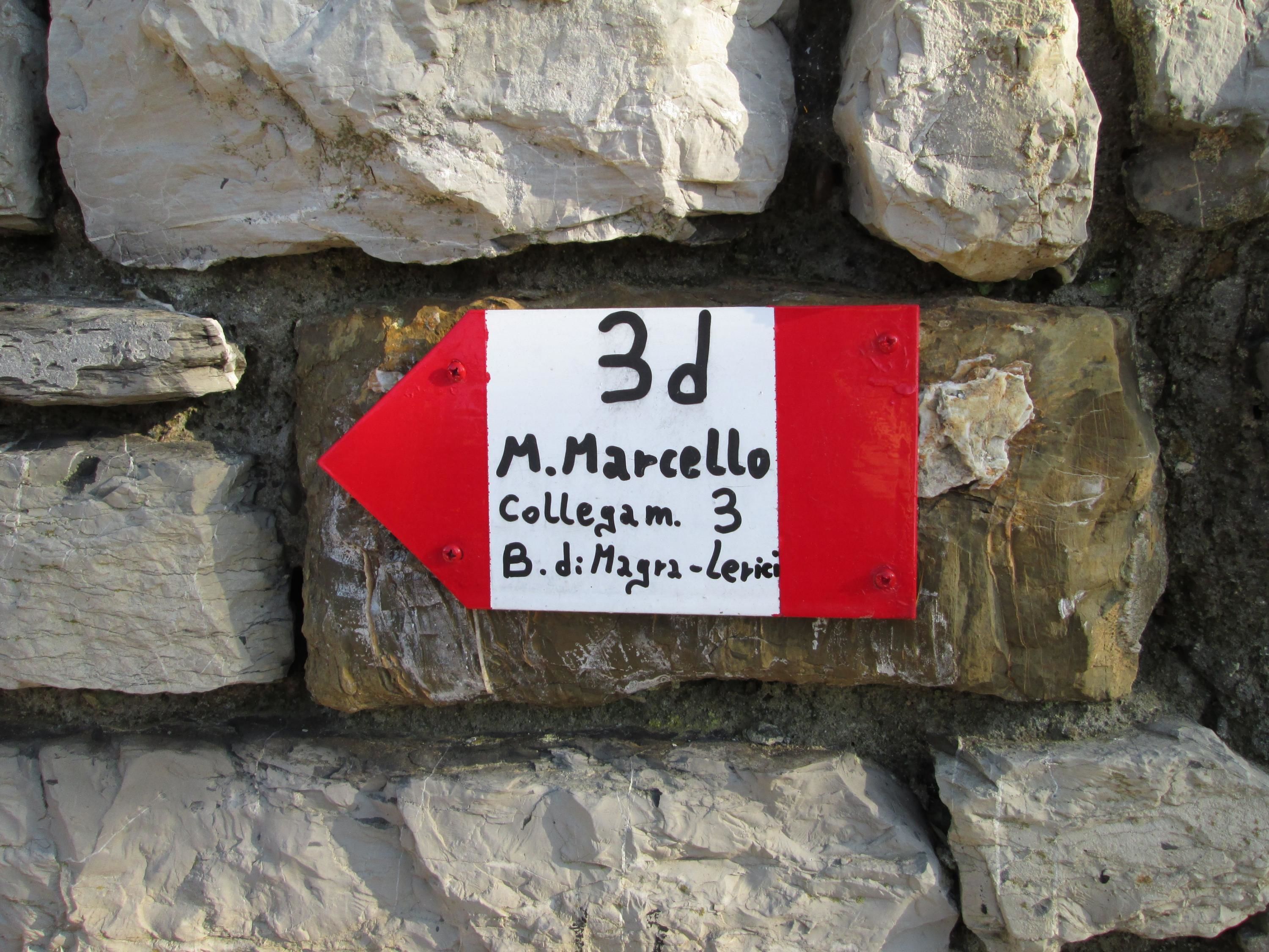 A directional sign on a stone wall, pointing to the left with the text '3d M. Marcello Collegam. 3 B. d. Magna-Lerici'.