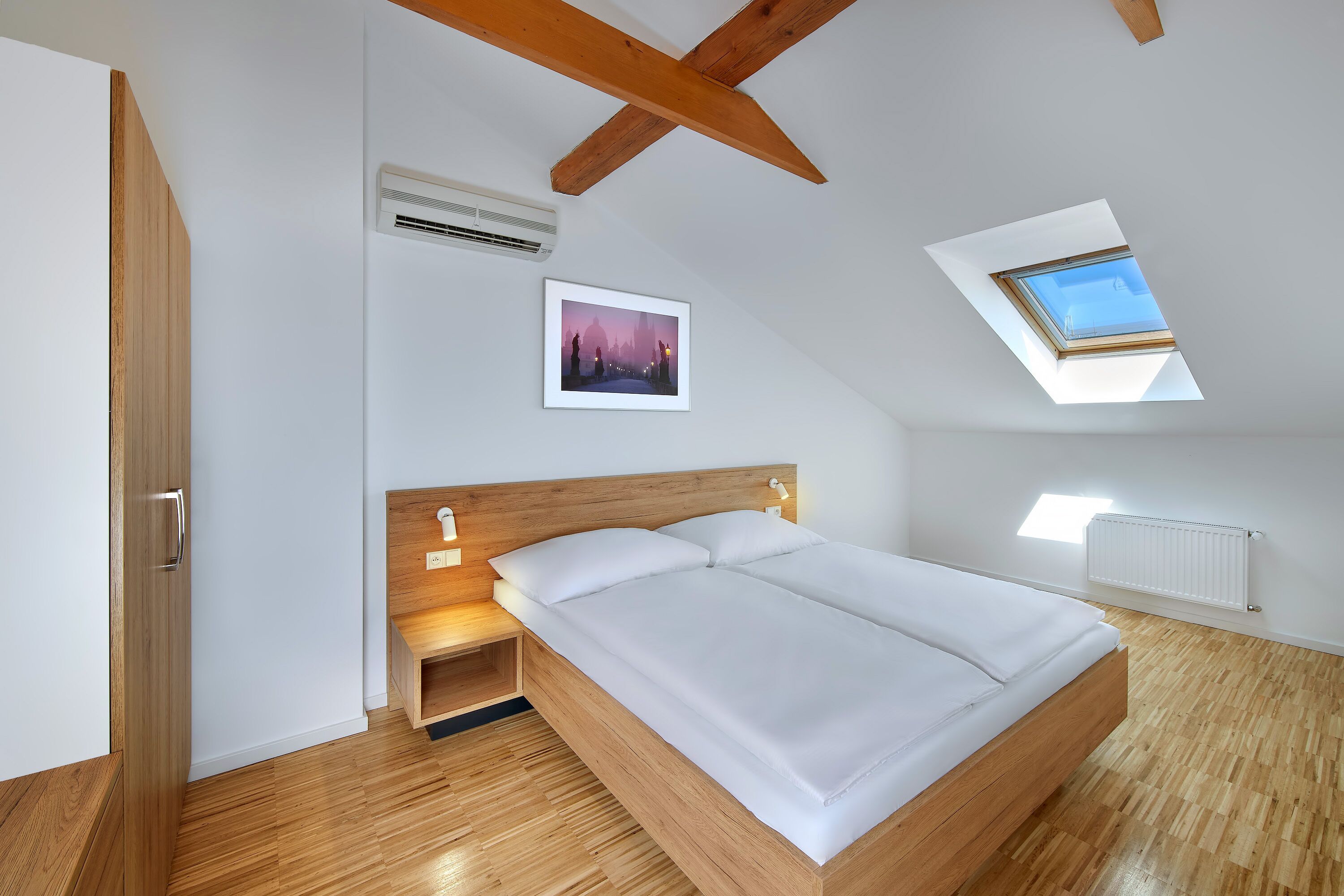 Un dormitorio con techo inclinado, vigas de madera, tragaluces, una cama, una mesita de noche y aire acondicionado instalado en la pared.